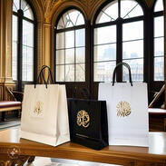 Cream and black shopping bags with gold foil logos placed on an ornate bench by an arched window with city views.